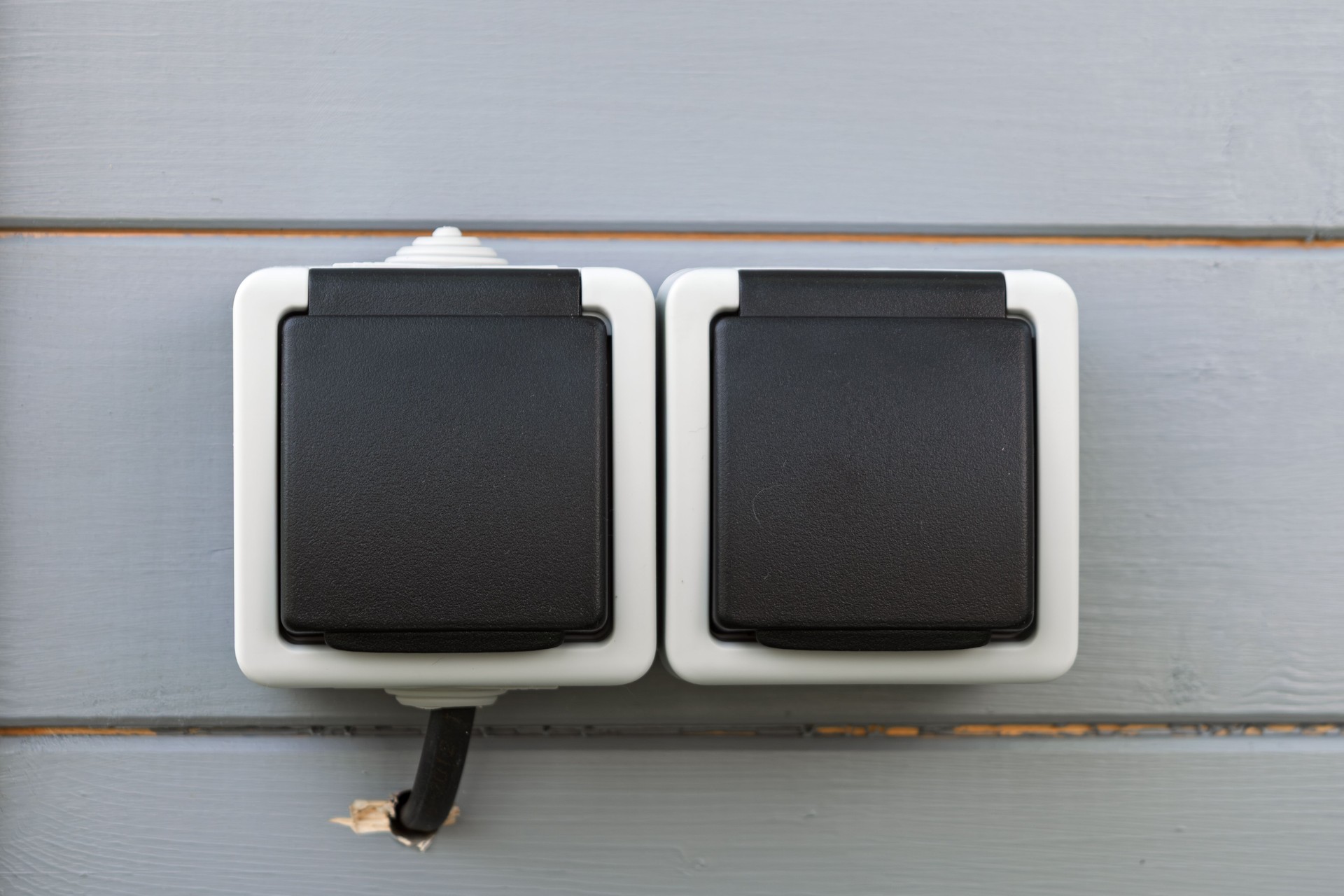 Modern double outdoor socket with closed black covers mounted on gray wooden wall offers weatherproof power access and clean design.